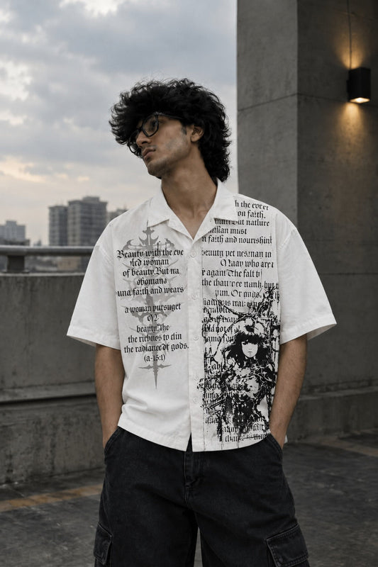 A man wearing a white short-sleeve button-up shirt with black graphic text and print on the front, paired with dark cargo pants, standing outdoors against a concrete wall.