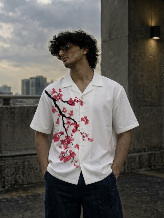 A man wearing a white short-sleeve button-up shirt with a red and black cherry blossom print, standing outdoors against a concrete wall and cloudy sky.