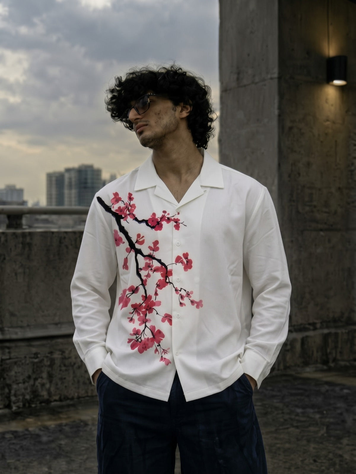 A man wearing a white long-sleeve button-up shirt with a pink cherry blossom branch print on the front, paired with dark trousers, standing outdoors against a concrete wall and cloudy sky.