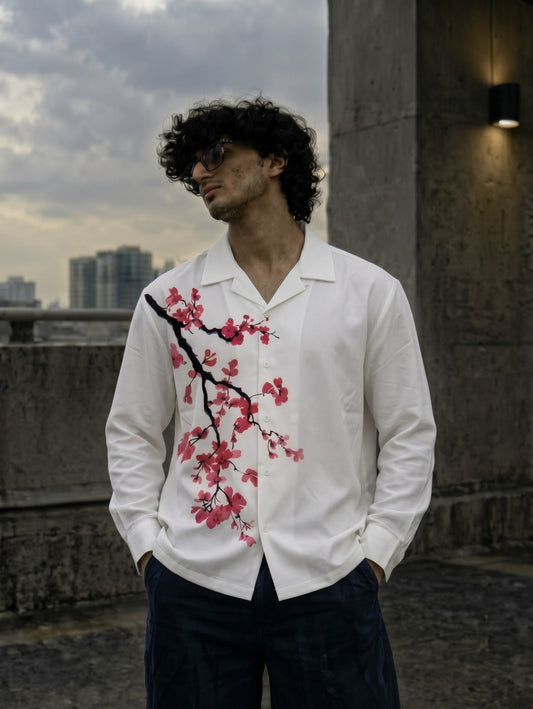 A man wearing a white long-sleeve button-up shirt with a pink cherry blossom branch print on the front, paired with dark trousers, standing outdoors against a concrete wall and cloudy sky.