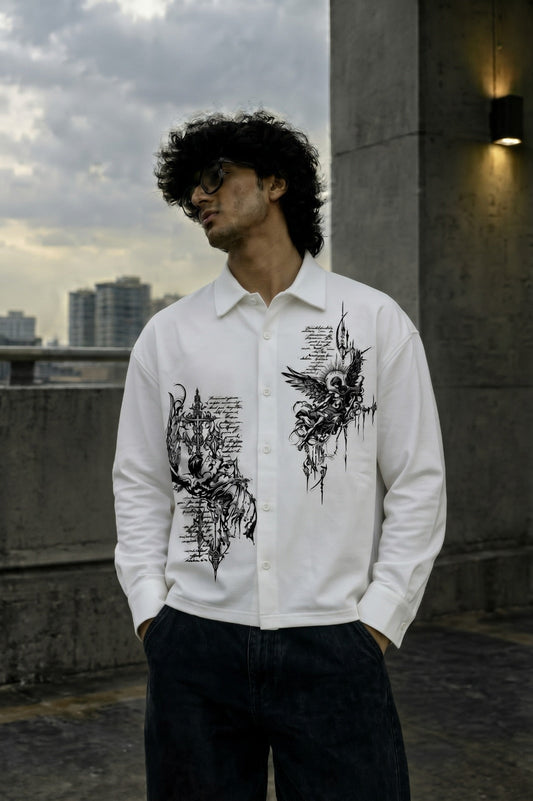 A man wearing a white long-sleeve button-up shirt with black graphic prints on the front, paired with dark jeans, standing outdoors on a concrete rooftop with a cloudy sky and city buildings in the background.