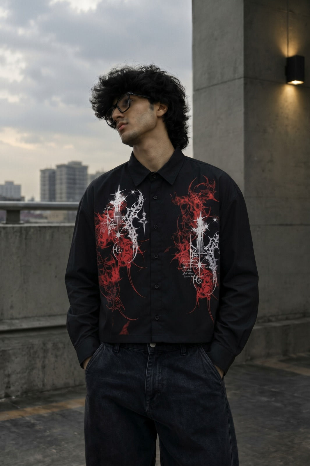 A man wearing a black long-sleeve button-up shirt with red and white graphic prints on the front, paired with dark jeans, standing on a concrete rooftop with a city skyline in the background.