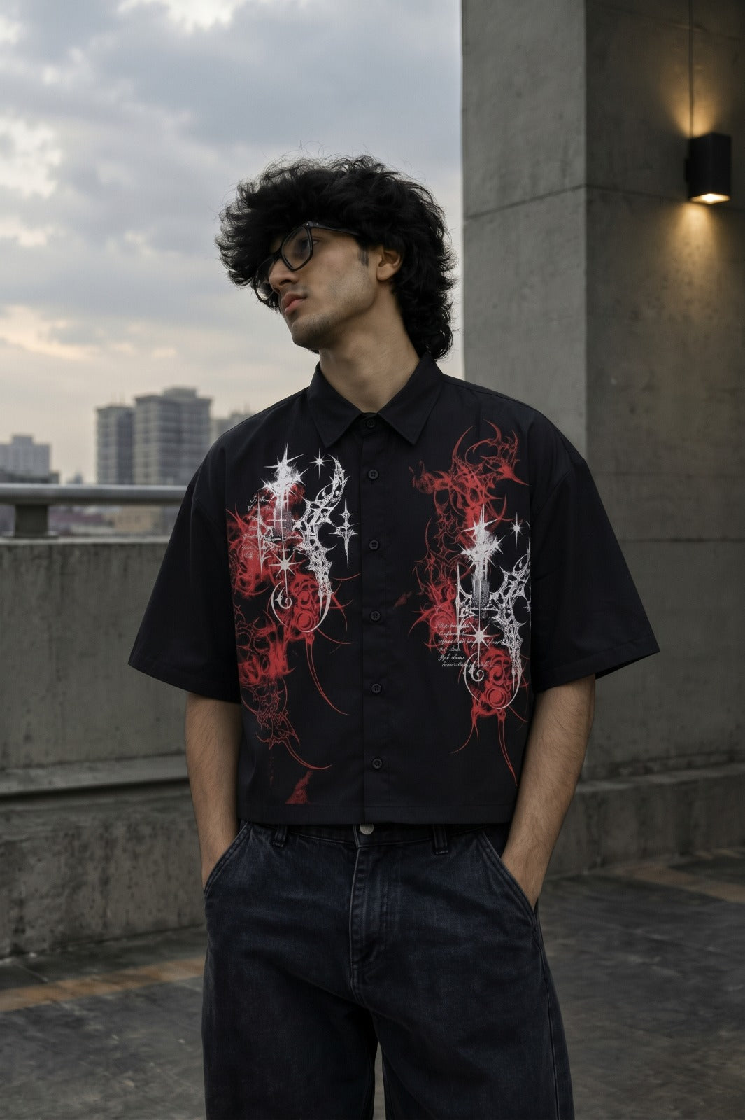 A man wearing a black short-sleeve button-up shirt with red and white graphic prints, standing on an outdoor rooftop with a city skyline in the background.
