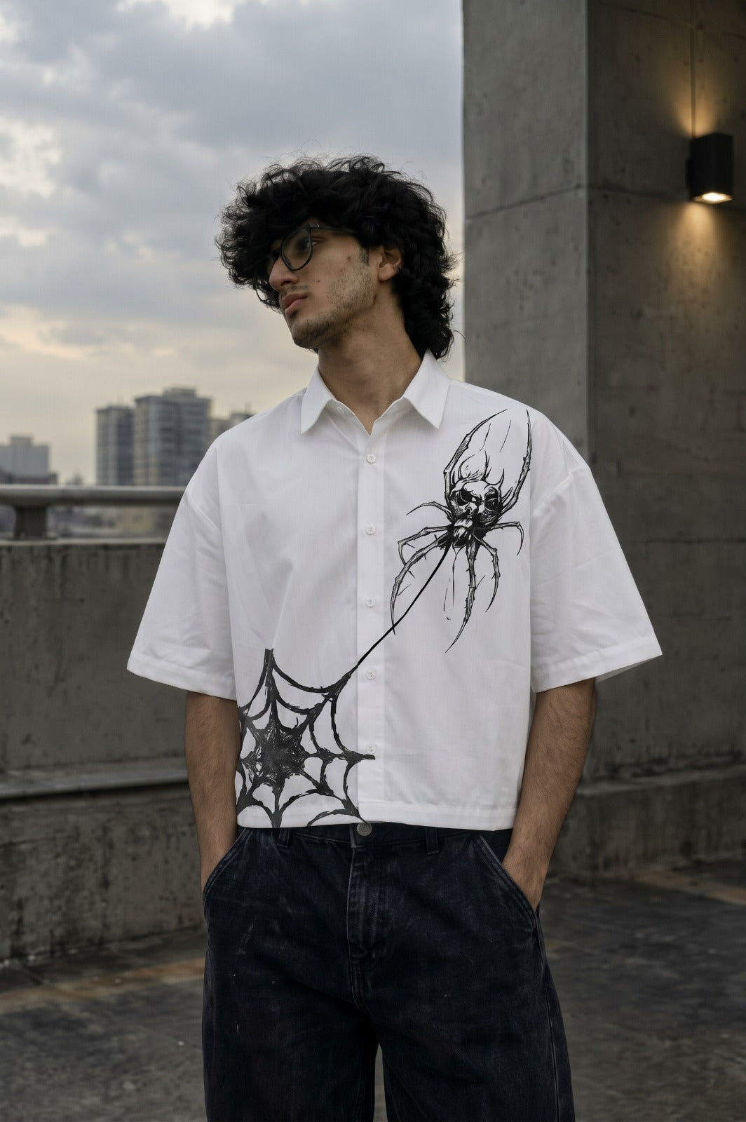 A man wearing a white short-sleeve button-up shirt with black spiderweb and spider graphics, paired with dark jeans, standing outdoors on a rooftop with a cloudy sky and city buildings in the background.