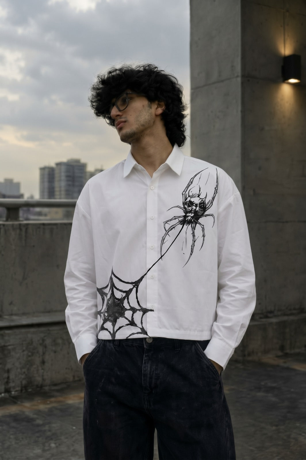 A man wearing a white long-sleeve button-up shirt with a black spider and web graphic print, paired with dark jeans, standing outdoors against a concrete wall and cloudy sky.