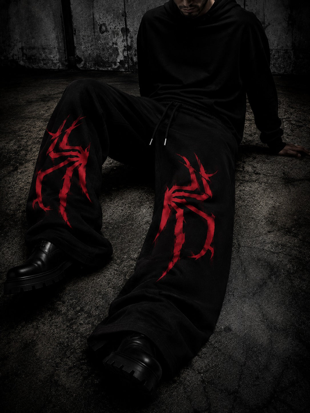 A person wearing black sweatpants with a large red spider graphic on each leg, seated on a dark concrete floor against a textured wall.