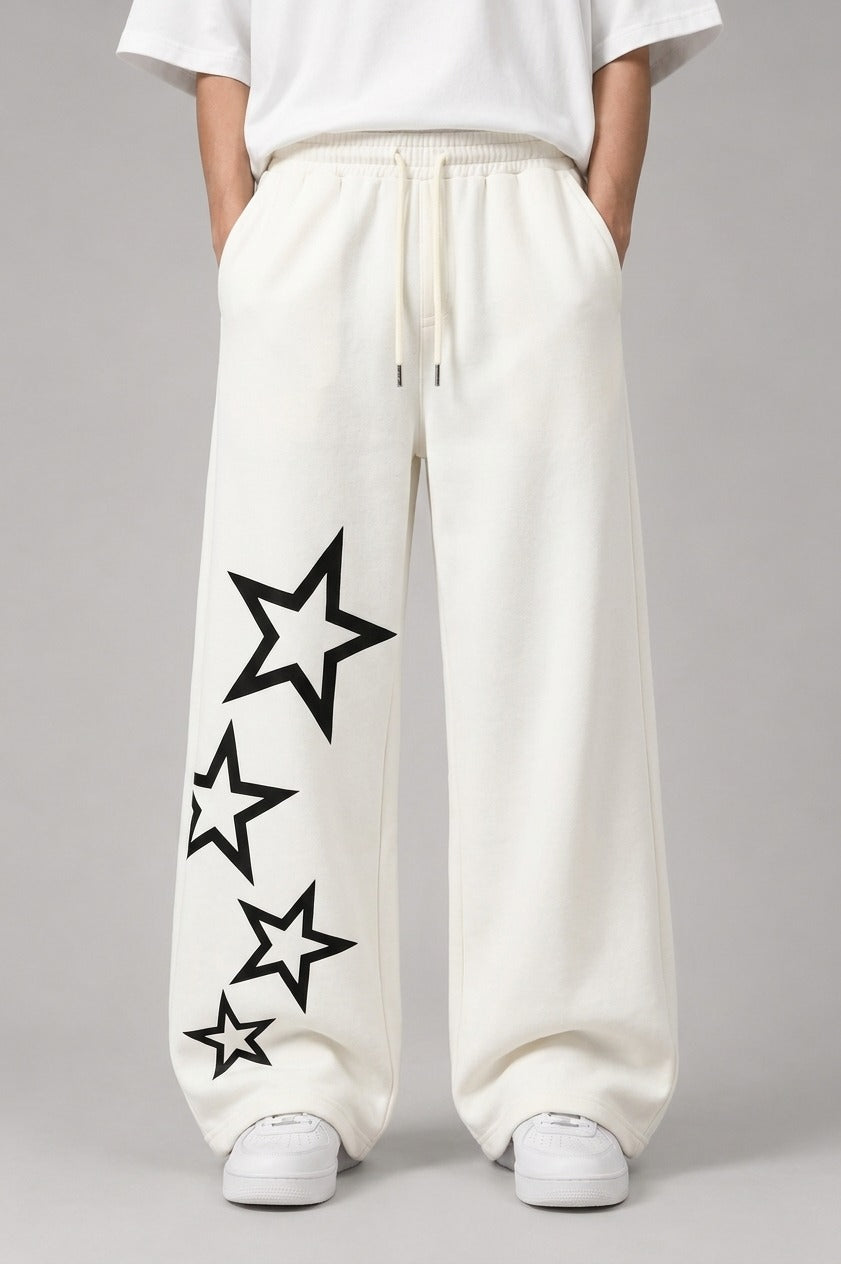 A person wearing loose white drawstring sweatpants with large black outlined star graphics printed down one leg, paired with a white top and white sneakers, against a light gray studio background.
