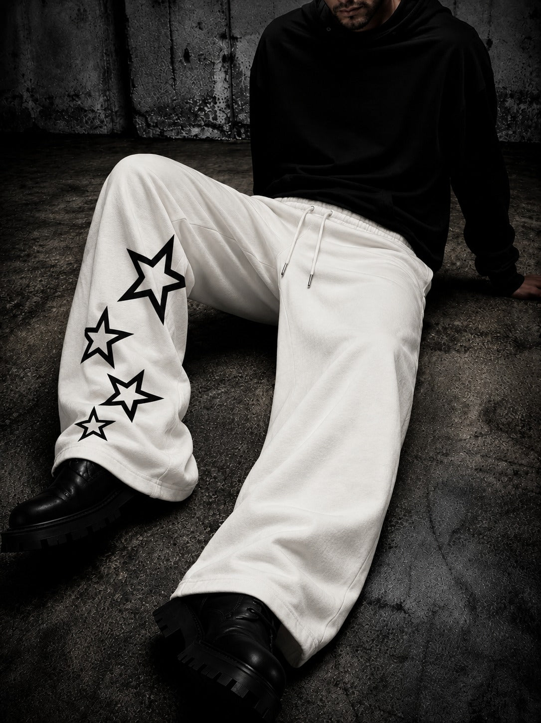A person sitting on a concrete floor wearing white wide-leg sweatpants with black star graphics on one leg, a black hoodie, and black boots.