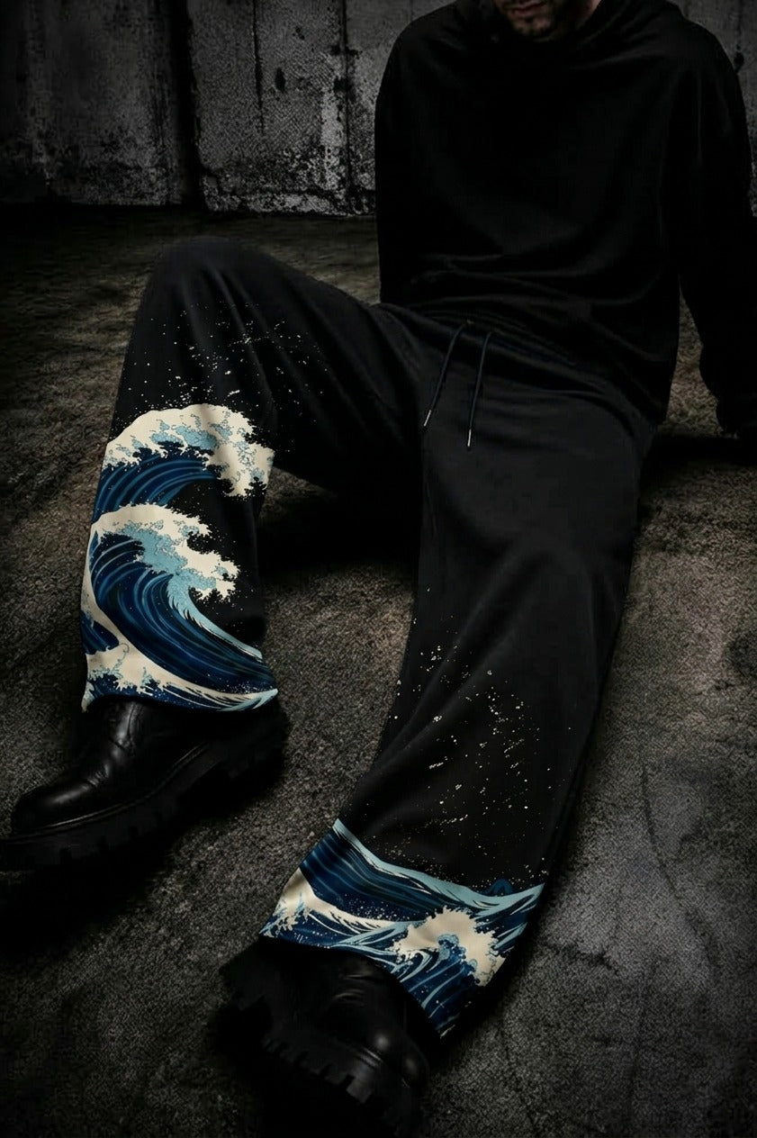 A person seated on a dark concrete floor wearing black sweatpants with a blue and white wave print at the lower legs, paired with black boots and a black hoodie.