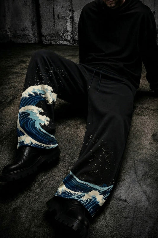 A person seated on a dark concrete floor wearing black sweatpants with a blue and white wave print at the lower legs, paired with black boots and a black hoodie.