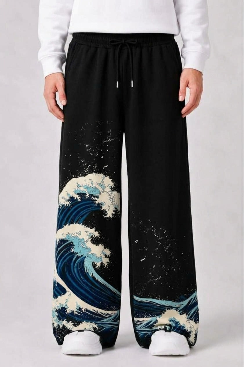 Black wide-leg sweatpants featuring a blue and white wave print inspired by 'The Great Wave off Kanagawa' on the lower legs, shown on a model wearing a white sweatshirt and white sneakers.