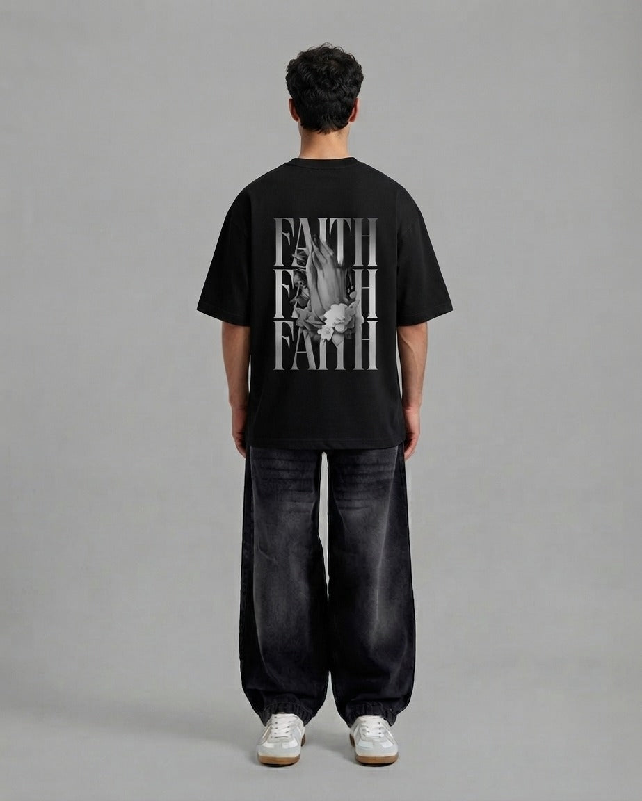A man viewed from behind wearing a black t-shirt with a large white graphic print on the back, paired with dark wide-leg jeans and white sneakers, against a solid gray studio background.