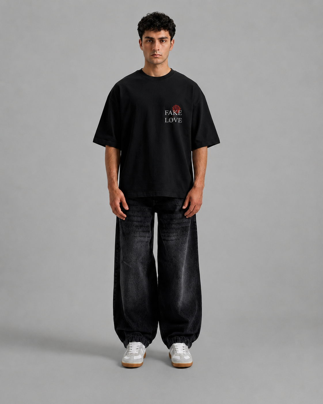 A man stands against a plain gray studio background wearing an oversized black T-shirt with a small red and white chest graphic reading 'FAKE LOVE', loose black jeans, and white sneakers.