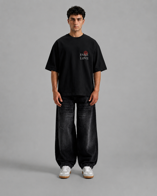 A man stands against a plain gray studio background wearing an oversized black T-shirt with a small red and white chest graphic reading 'FAKE LOVE', loose black jeans, and white sneakers.