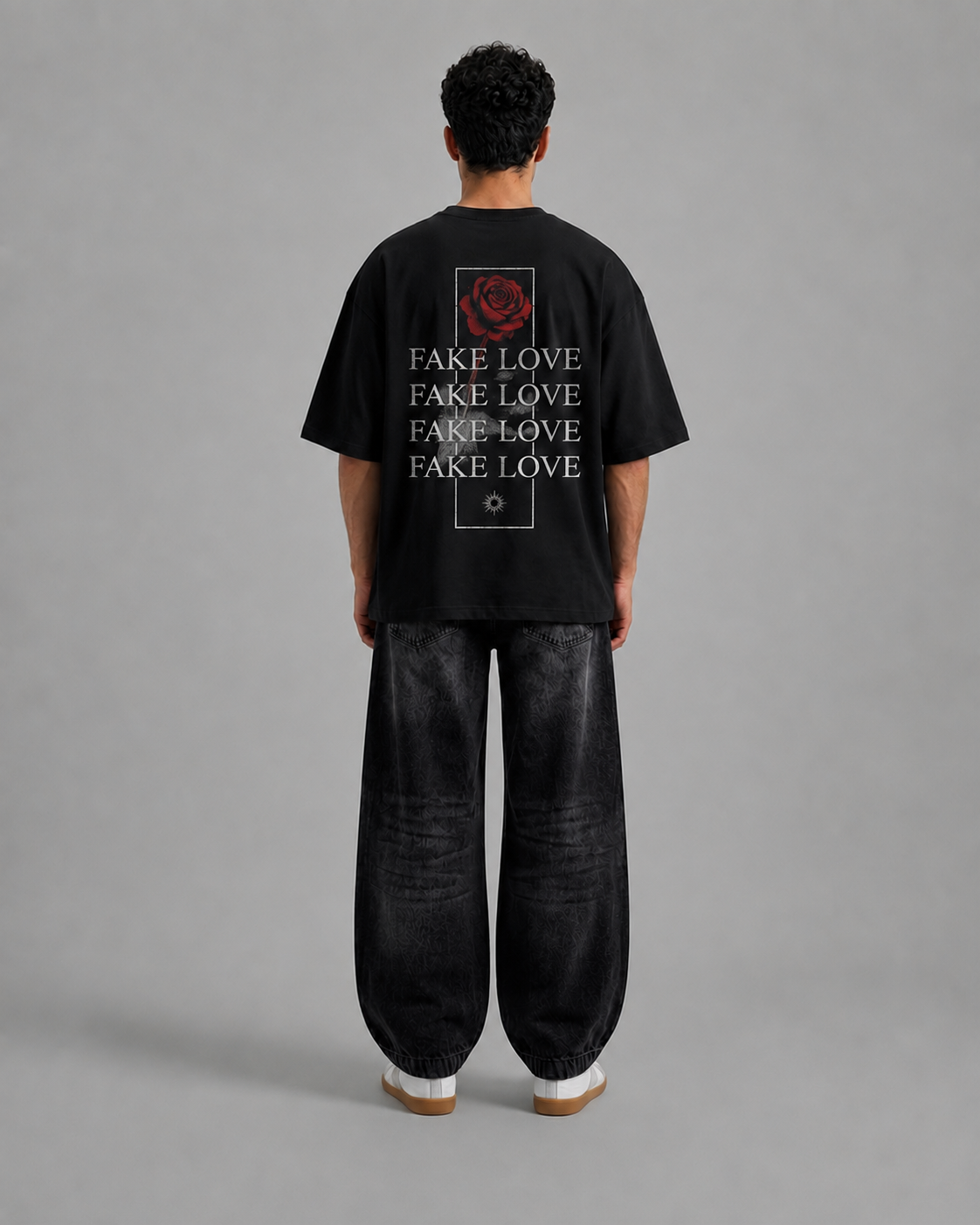 A man viewed from behind wearing a black oversized t-shirt with a red rose graphic and the word 'FAKE LOVE' repeated vertically, paired with loose black pants and white sneakers, against a plain gray studio background.