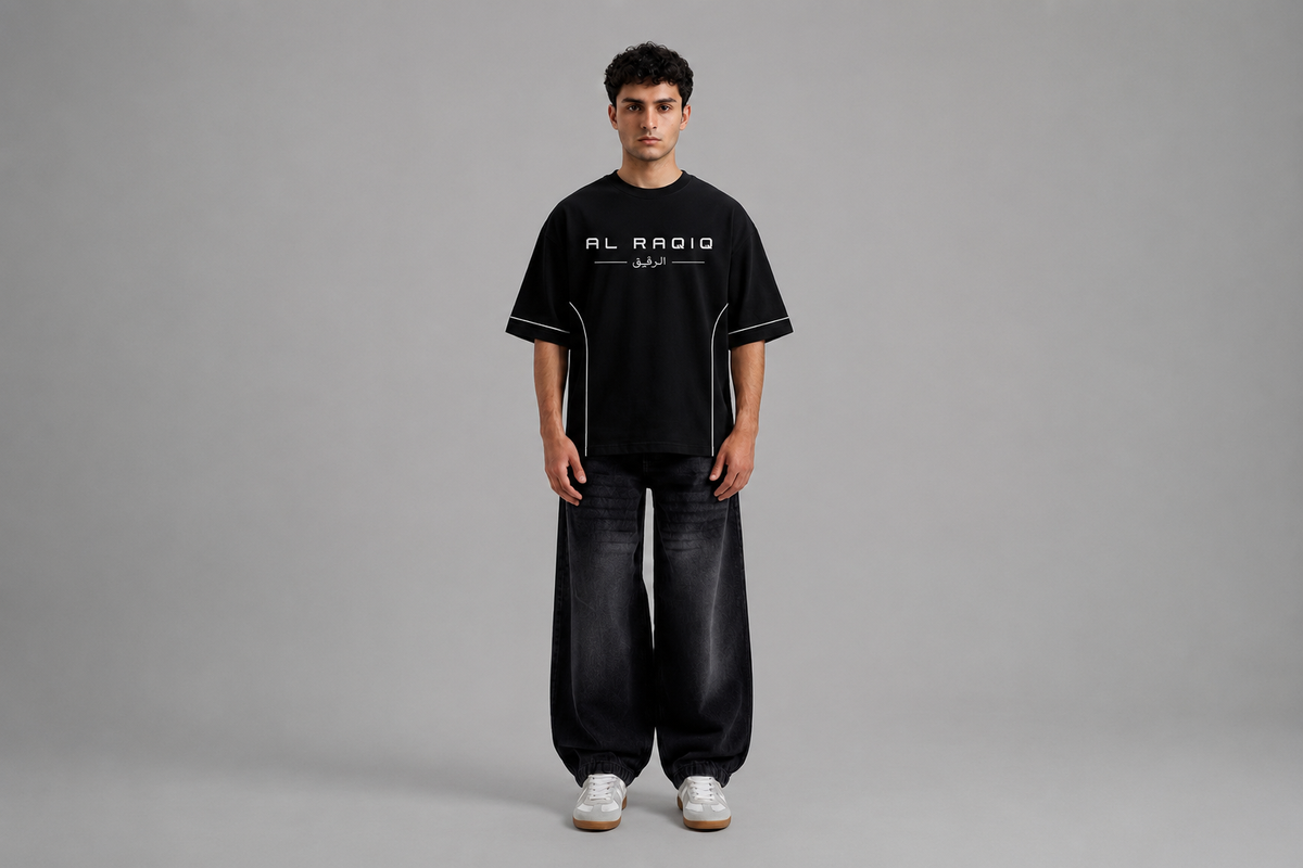 Load video: Male model showcasing a black oversized short-sleeve T-shirt with clean white piping details along the sleeves and sides, paired with dark wide-leg jeans in a minimalist studio setting.