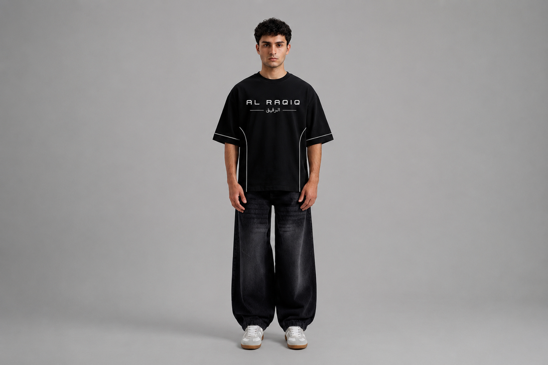Male model showcasing a black oversized short-sleeve T-shirt with clean white piping details along the sleeves and sides, paired with dark wide-leg jeans in a minimalist studio setting.