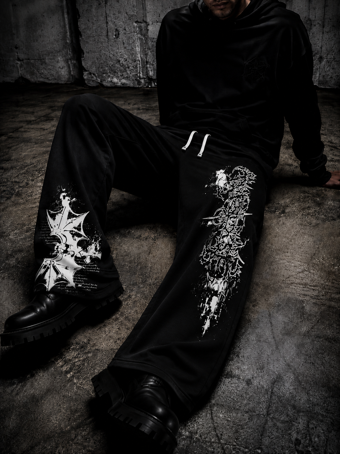 A person seated on a concrete floor wearing black wide-leg sweatpants with large white graphic prints on both legs, a black hoodie, and black boots in a dark industrial setting.