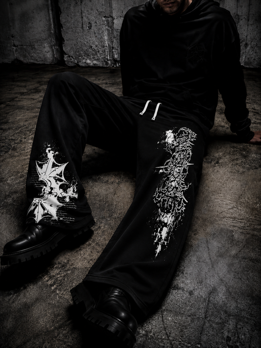 A person seated on a concrete floor wearing black wide-leg sweatpants with large white graphic prints on both legs, a black hoodie, and black boots in a dark industrial setting.