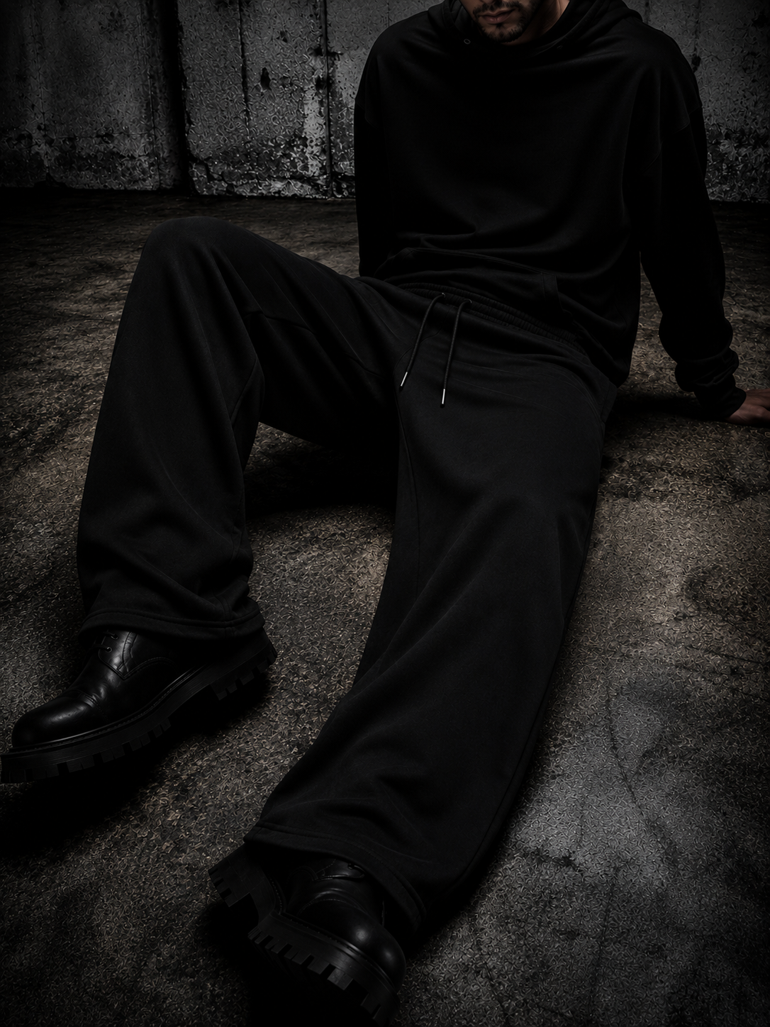 A person wearing a black hoodie and black wide-leg sweatpants sits on a textured concrete floor against a dark wall.