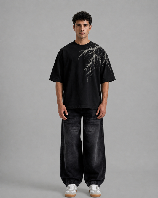 A man modeling a black short-sleeve t-shirt with a white branch graphic on the chest, paired with baggy dark jeans and white sneakers, against a solid gray studio background.
