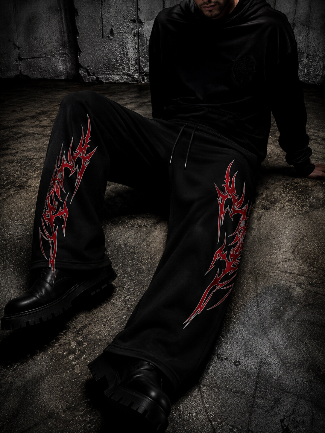 A person wearing black sweatpants with large red and white tribal-style graphic prints on both legs, seated on a concrete floor against a dark textured wall.