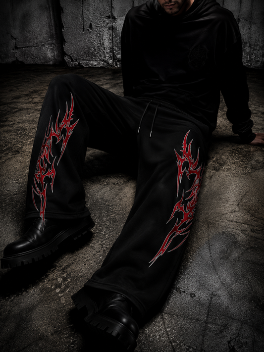 A person wearing black sweatpants with large red and white tribal-style graphic prints on both legs, seated on a concrete floor against a dark textured wall.