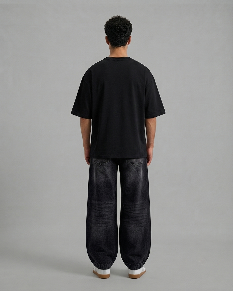 A man viewed from behind wearing a plain black short-sleeve t-shirt and very baggy dark charcoal gray pants, standing against a solid light gray studio background.