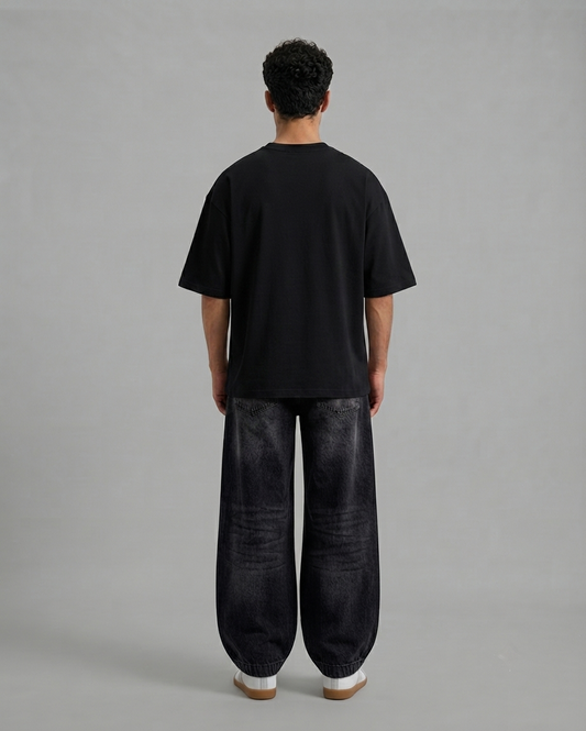 A man viewed from behind wearing a plain black short-sleeve t-shirt and very baggy dark charcoal gray pants, standing against a solid light gray studio background.