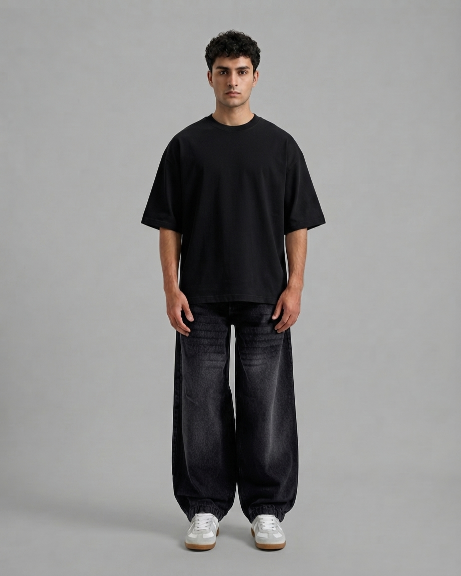 A man stands against a plain light gray studio background wearing an oversized black short-sleeve T-shirt, loose dark charcoal pants, and white sneakers.