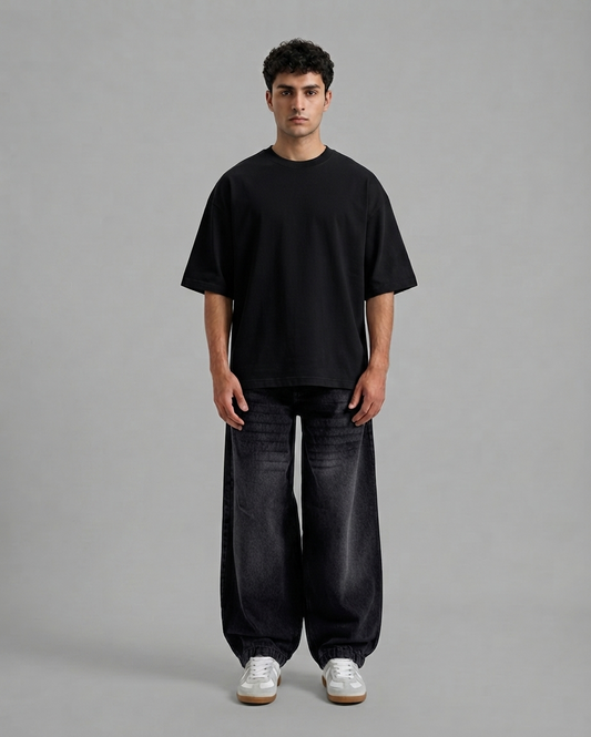A man stands against a plain light gray studio background wearing an oversized black short-sleeve T-shirt, loose dark charcoal pants, and white sneakers.